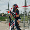 Person in lacrosse gear standing next to a goalpost with a red ball on a field.