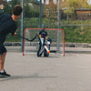 Two people playing lacrosse on an outdoor court with a goal in the background.